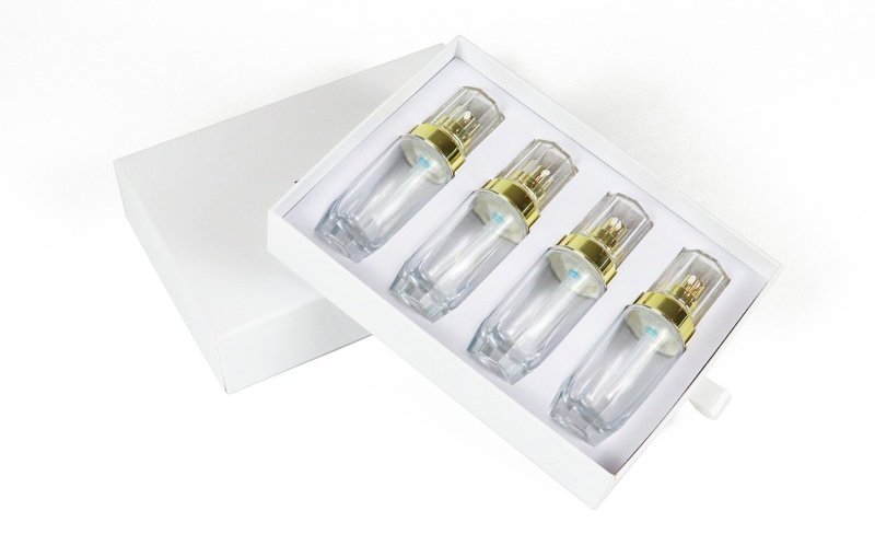 White card beauty gift box (open) containing 4 high-end clear cosmetic bottles (gold-accented pumps) in tailored inner cushioning